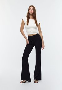 Bela kratka majica s kratkimi rokavi, v kombinaciji s črnimi širokimi hlačami. Manekenka nosi črne sandale na blok peto; gladka tekstura tkanine.