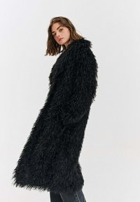 Černý kabát z umělé kožešiny s dlouhým, volným střihem, texturovaným povrchem a plnými rukávy. Nošený přes tmavé džíny na neutrálním pozadí.