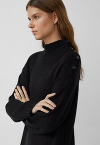 Zwarte trui met hoge nek voorzien van ribbelstructuur en decoratieve knopen langs de linkerschouder. Lange mouwen en een ontspannen pasvorm.