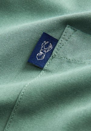Groen textiel met een gladde textuur en een marineblauw label, met daarop een wit geborduurd ontwerp van hertenafwerpen.