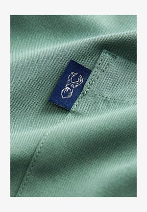 Material textil verde cu o textură netedă, având o etichetă albastru închis cu un design brodat alb de coarne.