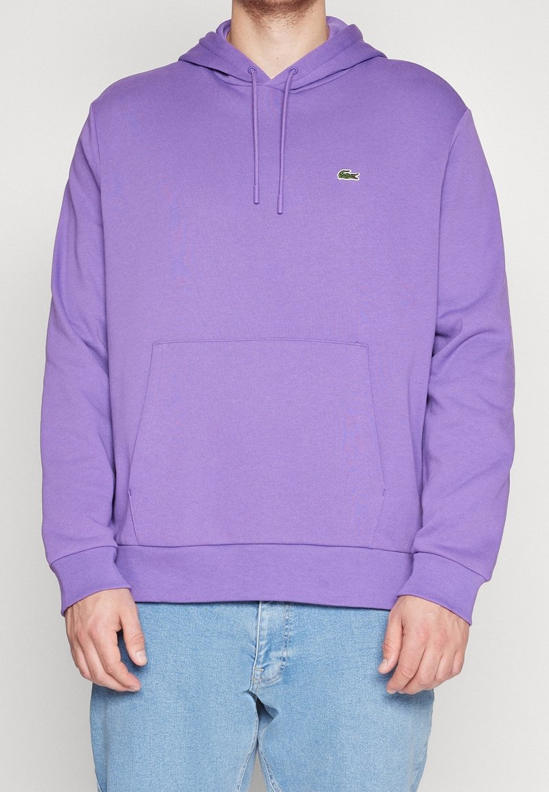 Homme portant un sweat à capuche lavande avec poche avant et cordons, associé à un jean bleu clair, sur un fond uni.