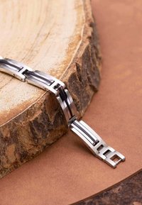 Metalen horlogearmband met afwisselende zilveren en zwarte schakels. Heeft een gepolijste afwerking en een veilige sluiting, geplaatst op houten en leren oppervlakken.