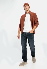 Brązowy sweter z teksturą na zamek, biały t-shirt, ciemne dżinsy i jasno szare sneakersy. Model pozujący na neutralnym tle.