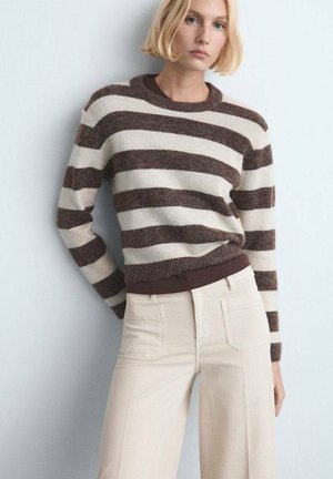 Mujer con melena rubia estilo bob que lleva un suéter marrón con rayas blancas, metido dentro de unos pantalones amplios de tiro alto color crema, de pie frente a un fondo claro.