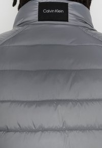 Grau gefütterte Jacke mit gesteppter Optik, ausgestattet mit einem hohen Kragen und einem schwarzen Etikett mit weißem Text auf dem Rücken. Glatte, glänzende Textur.