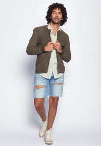 Olivgrüne Bomberjacke, blumiges Button-Up-Hemd, zerfetzte Jeansshorts und weiße Sneaker. Ein lässiges, geschichtetes Outfit wird präsentiert.