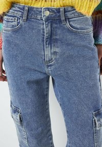 Jeans de mezclilla azul de tiro alto con un lavado desvanecido, bolsillos estándar y un bolsillo de carga en el muslo izquierdo. Tejido de mezclilla texturizado.