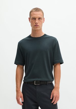 Jonge man met kort haar, die een donkergroen shirt met korte mouwen en een zwarte broek met zwarte riem draagt, staande tegen een effen witte achtergrond.
