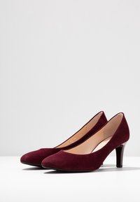 Ett par mörkröda mocka pumps med mellanlång klack och spetsiga tår på en vit yta mot en ljusgrå bakgrund.