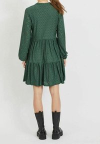 Grön långärmad klänning med ett strukturmönster som liknar spets, flerlagig kjol och en figursydd överdel. Bärs med svarta knähöga stövlar.