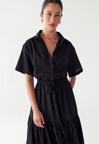 Juoda lininė suknelė su trumpomis rankovėmis, mygtukiniu užsegimu ir elastingu juosmeniu. Pasižymi sluoksniuota sijona ir reguliuojamu raišteliu.