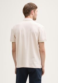 Beige Poloshirt mit kurzen Ärmeln und weicher Textur, klassischem Kragen und grünen Akzenten an den Ärmeln. Nur Rückansicht.