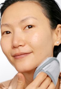 Un tampon de nettoyage du visage avec une texture en jersey grise et douce, maintenu contre la mâchoire, avec un léger éclat sur la peau et des imperfections minimales.