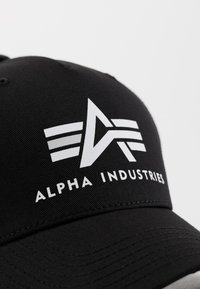 Fekete baseball sapka, elöl egy fehér logóval, három csíkkal és "ALPHA INDUSTRIES" felirattal. Simább szövettextúra.