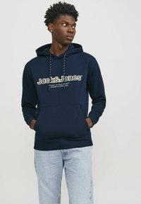 Sudadera azul marino con bolsillo frontal, capucha con cordones y texto del logo. Tela suave con puños y dobladillo acanalado, combinada con jeans azul claro.