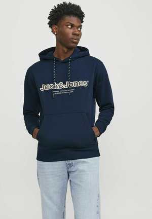 Marineblaue Kapuzenjacke mit Fronttasche, Kordelzugkapuze und Logo-Text. Aus weichem Stoff mit gerippten Bündchen und Saum, getragen mit hellblauen Jeans.