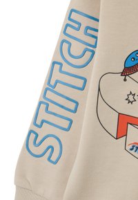 Felpa beige con testo blu "STITCH", caratterizzata da un design grafico di un personaggio con un cappello. Tessuto morbido, vestibilità standard, polsini a costine.