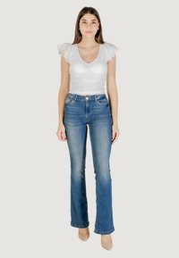 Top bianco a coste con dettagli in pizzo, abbinato a jeans a zampa d'elefante blu. Modella in piedi, indossa tacchi beige. Sfondo neutro.