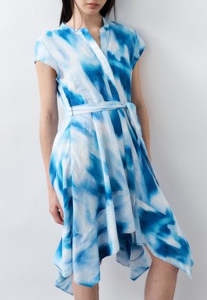 Naine, kes kannab sinise ja valge lipsuvärviga vööga kleiti koos kapuutsvarrukate ja asümmeetrilise hemegaraažiga, seisab tavalise seina ees.