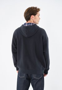 Sudadera negra con capucha, hecha de una tela suave, que presenta un corte relajado, puños de canalé y una camisa de cuadros en capas visible en el cuello.