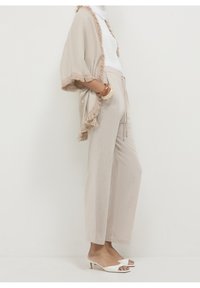 Licht beige kimono met franjes, gecombineerd met loszittende licht beige broek. Witte open tenen sandalen maken de outfit compleet.