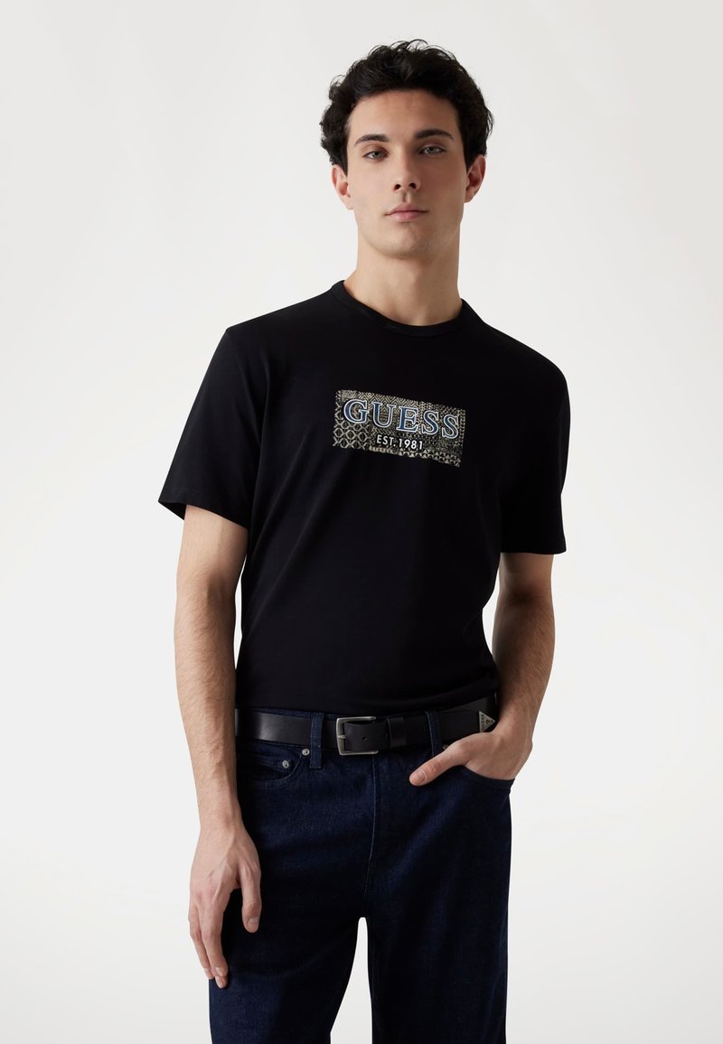 Czarny t-shirt z teksturowanym nadrukiem „GUESS EST. 1981” w srebrnym i białym kolorze. W połączeniu z ciemnoniebieskimi dżinsami i czarnym paskiem.