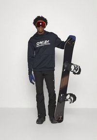 Σνόουνμπορντ με ναυτικό φούτερ με λευκό λογότυπο, μαύρα παντελόνια σκι, μπλε γάντια και γυαλιά, κρατώντας έναν μαύρο σνόουνμπορντ με λευκά δεσίματα.