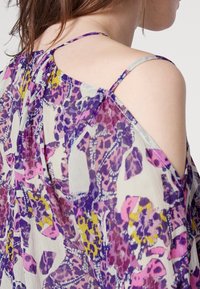 Femme portant un top épaules dénudées avec fines bretelles, présentant un motif floral et abstrait coloré en violet, rose, jaune et blanc.