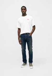 Λευκό κοντομάνικο μπλουζάκι με κόκκινο λογότυπο «DIESEL», σε συνδυασμό με σκούρο μπλε τζιν παντελόνι και γκρι αθλητικά παπούτσια, στέκεται μπροστά σε ουδέτερο φόντο.