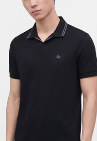 Homme portant un polo noir avec un petit logo A|X et des détails de col rayés gris sur un fond clair uni.