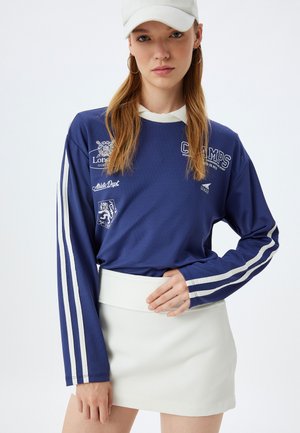 Long-sleeve navy shirt with white stripes and logos, paired with a white skirt. The shirt features a textured fabric.