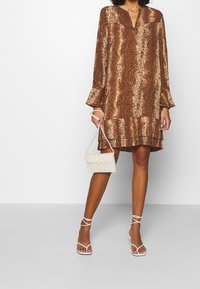 Braun gemustertes Kleid mit langen Ärmeln, V-Ausschnitt und gestuftem Saum. Ergänzt durch eine kleine weiße Handtasche und weiße Sandalen mit Riemchen.