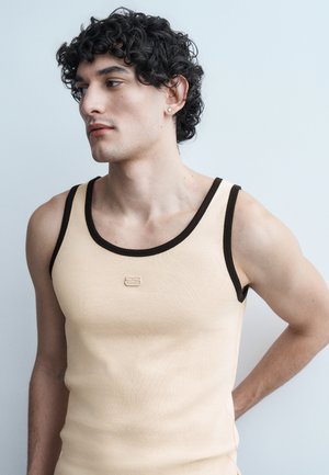 Νέος άνδρας με σγουρά μαύρα μαλλιά, φορώντας μπεζ φανέλα χωρίς μανίκια με μαύρες λεπτομέρειες, στέκεται μπροστά σε ανοιχτό γαλάζιο φόντο, κοιτάζοντας προς τα αριστερά.