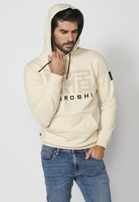 Sudadera beige con un bolsillo canguro en la parte frontal, con cordones oscuros y texto impreso. Combinada con jeans azules, de textura de tela suave.