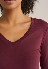 Bordo top s výstrihom do V s hladkou textúrou a priliehavým dizajnom, s klasickým strihom, ktorý zvýrazňuje oblasť goliera a ramien.