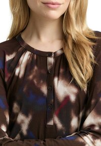 Braunes Langarmshirt mit rundem Ausschnitt, mit einer Knopfleiste und einem Batikmuster in Blau-, Rot- und Cremetönen.