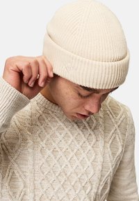 Beige neulepipo, jossa on ribbiohjelma ja pieni logo-tagi. Yhdistetty kevyenväriseen neulepaitaan, jossa on timanttikuvio.