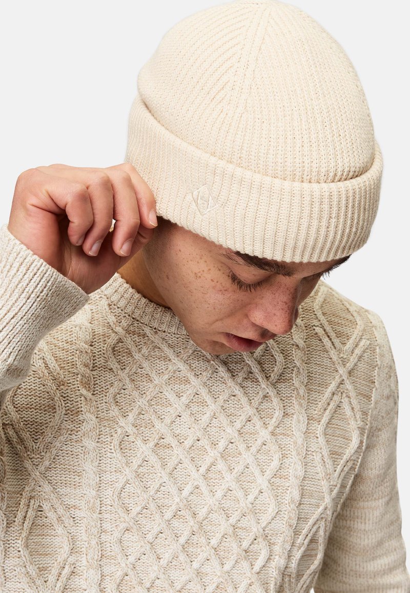 Beige neulepipo, jossa on ribbiohjelma ja pieni logo-tagi. Yhdistetty kevyenväriseen neulepaitaan, jossa on timanttikuvio.
