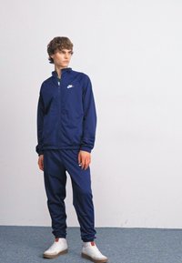 Námořnický velour set skládající se z hladké tkaniny, s bundou na zip a úzkými kalhotami, s bílým logem na horní levé straně.