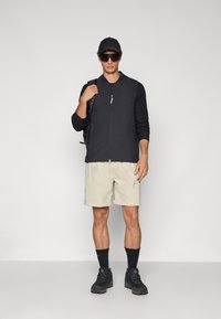 Black zip-up jacket with long sleeves, beige shorts, black socks, and outdoor shoes. Accessories include a black cap and sunglasses.