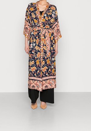 Femme portant une longue robe style kimono à motifs floraux bleu marine, pêche et orange, pantalon noir à jambes larges et escarpins couleur chair.