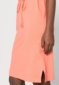 Vaaleanpunainen korallimekko, joka on valmistettu pehmeästä kankaasta, jossa on kiristettävä vyötärö ja koristeellisia kultaisia yksityiskohtia; hemuksessa on sivuaukot liikkuvuuden helpottamiseksi.