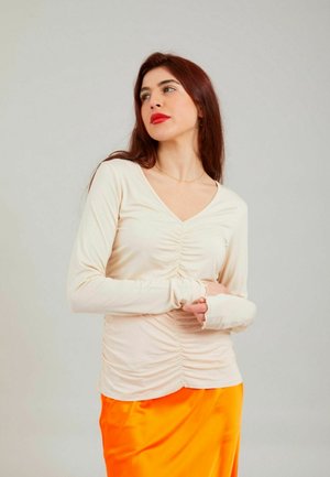 Frau mit langen roten Haaren und rotem Lippenstift, trägt ein cremefarbenes, gerafftes Langarmshirt und einen knallorangen Rock, blickt seitlich vor einem schlichten Hintergrund.