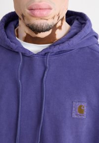 Sudadera con capucha morada de algodón, que cuenta con un bolsillo tipo canguro y un parche con el logo de Carhartt en la esquina superior izquierda.