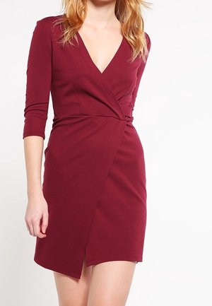 Mujer con un vestido cruzado burdeos ajustado hasta la rodilla, con mangas tres cuartos y escote en V.