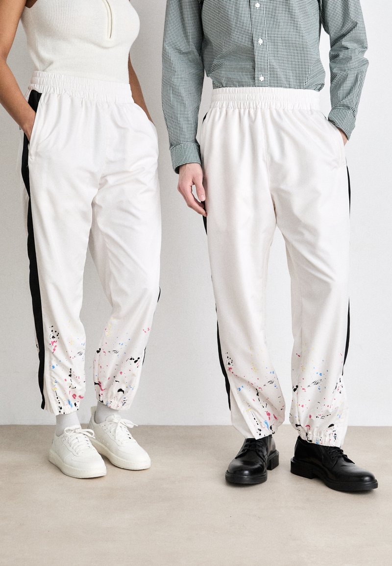 White athletic pants with black side panels, featuring colorful splatter patterns. Elastic waistband and cuffs, worn with white and black shoes.