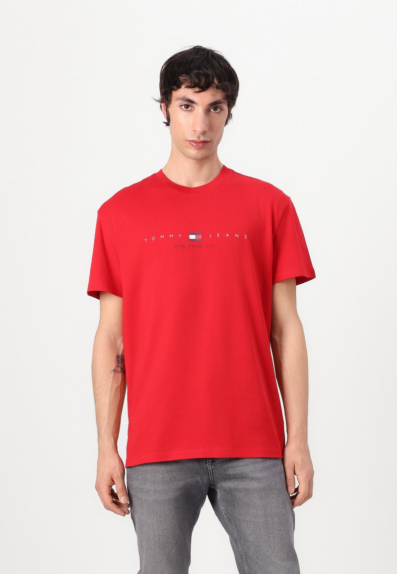 Crvena pamučna majica kratkih rukava s okruglim izrezom, s centralnim logotipom koji glasi "TOMMY JEANS" iznad male grafike zastave.