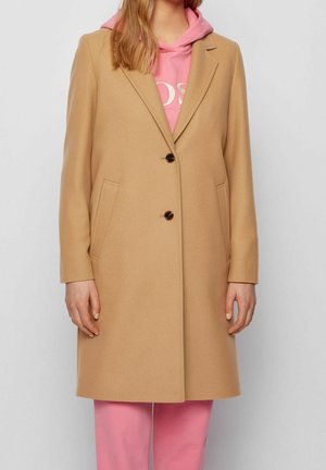Femme portant un manteau en laine couleur camel à deux boutons par-dessus un sweat à capuche rose et un pantalon assorti, debout devant un fond uni clair.