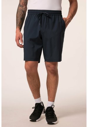 Mand iført sorte træningsshorts med snøre, hvide sokker og sorte sneakers, stående med den ene hånd i lommen.